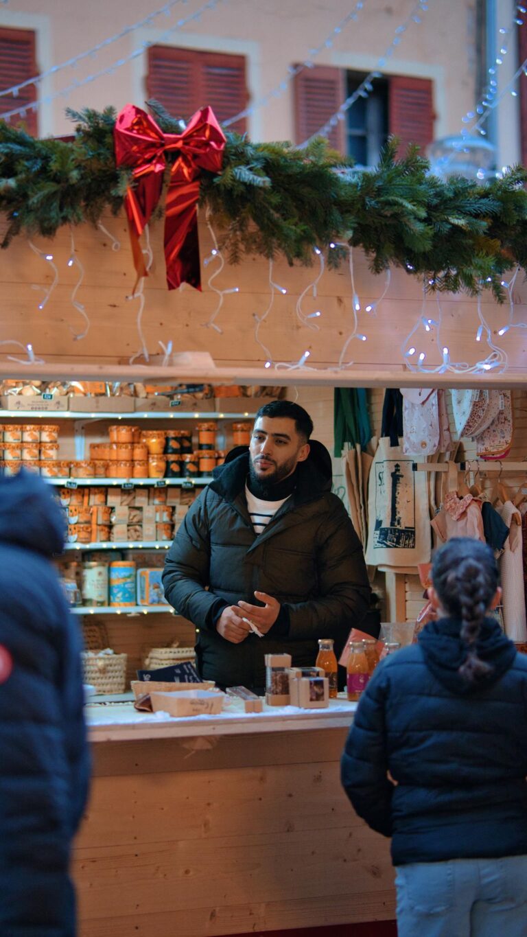 Man serving customers at a decorated Christmas market stall with festive lights.
